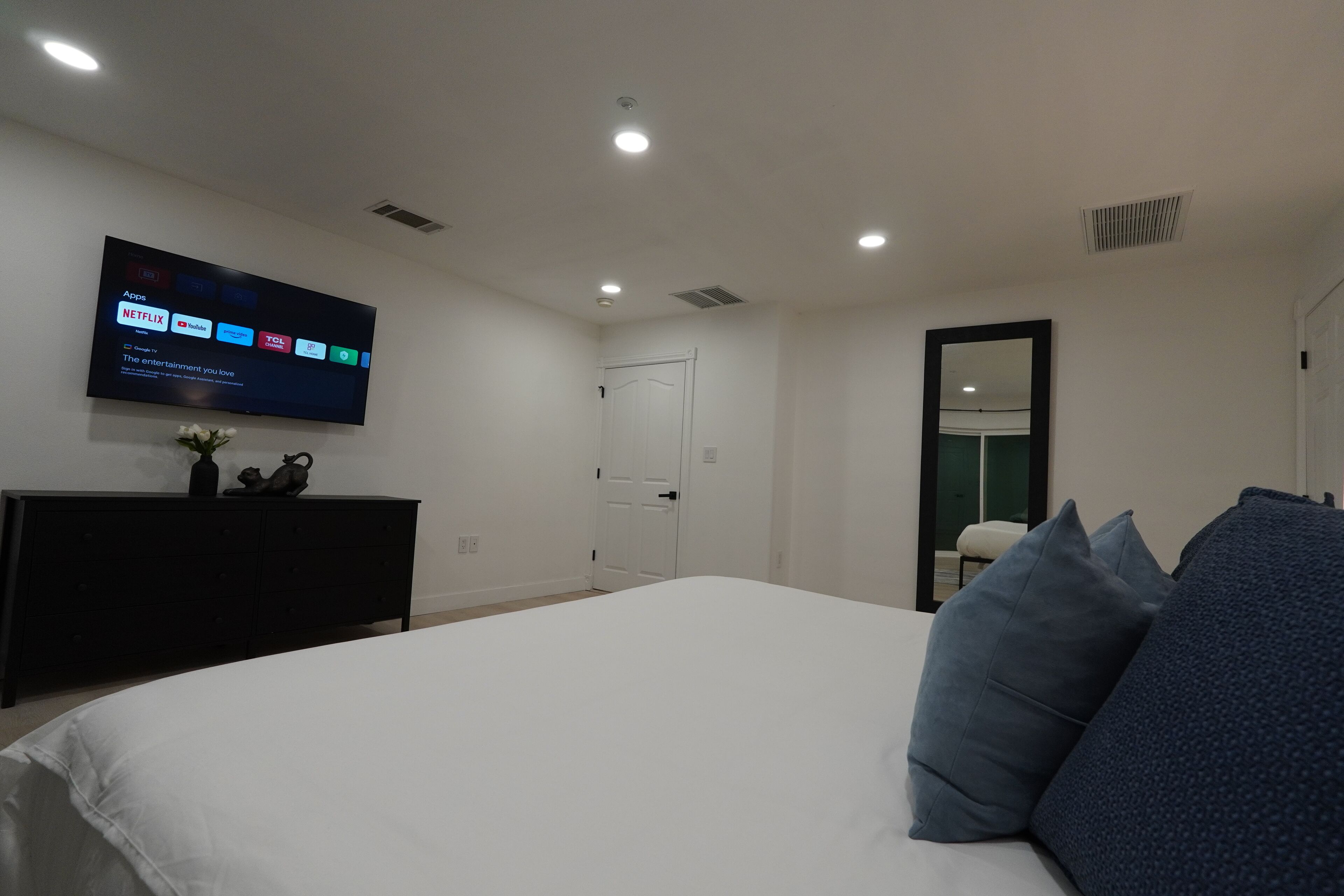 Un dormitorio moderno con una cama grande, un televisor de pantalla plana montado en la pared mostrando aplicaciones de transmisión, y un aparador oscuro.
