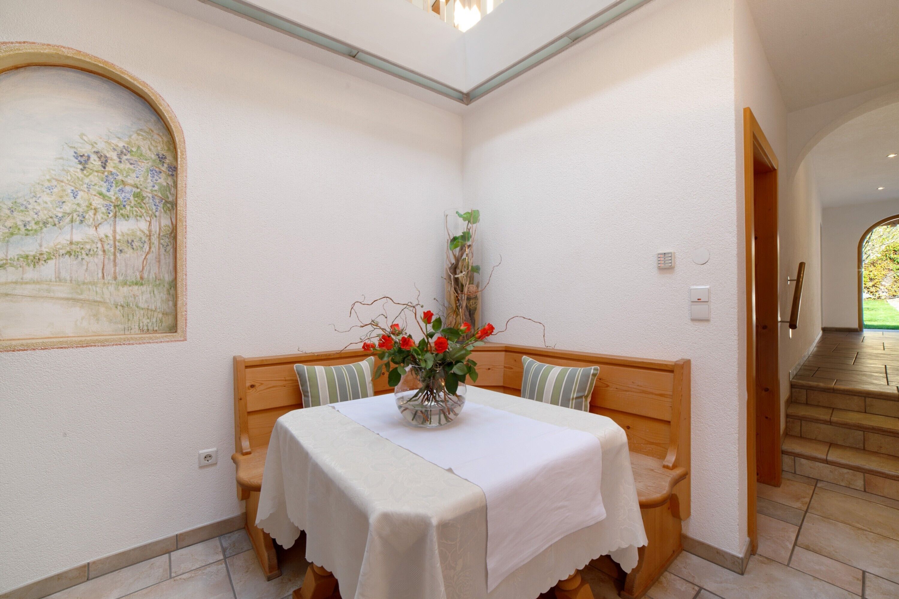 Un comedor con una mesa dispuesta para dos, un banco con cojines, un florero con flores y un cuadro en la pared.