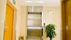 A modern elevator with a metallic finish, a wooden door on the left, and a potted plant on the right. - Anna Baige Hotel (Shanghai)