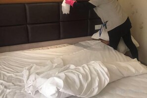 A bed with a white comforter and pillows, a headboard with a dark panel, and a person standing by the bed. - Anna Baige Hotel (Shanghai)