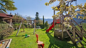 A playground with a slide and swings, surrounded by greenery and a mountain view. - Wiedmairhof Apartment 'Schwalbennest' with West-facing balcony, Shared Terrace & Garden and Wi-Fi (Lagundo)