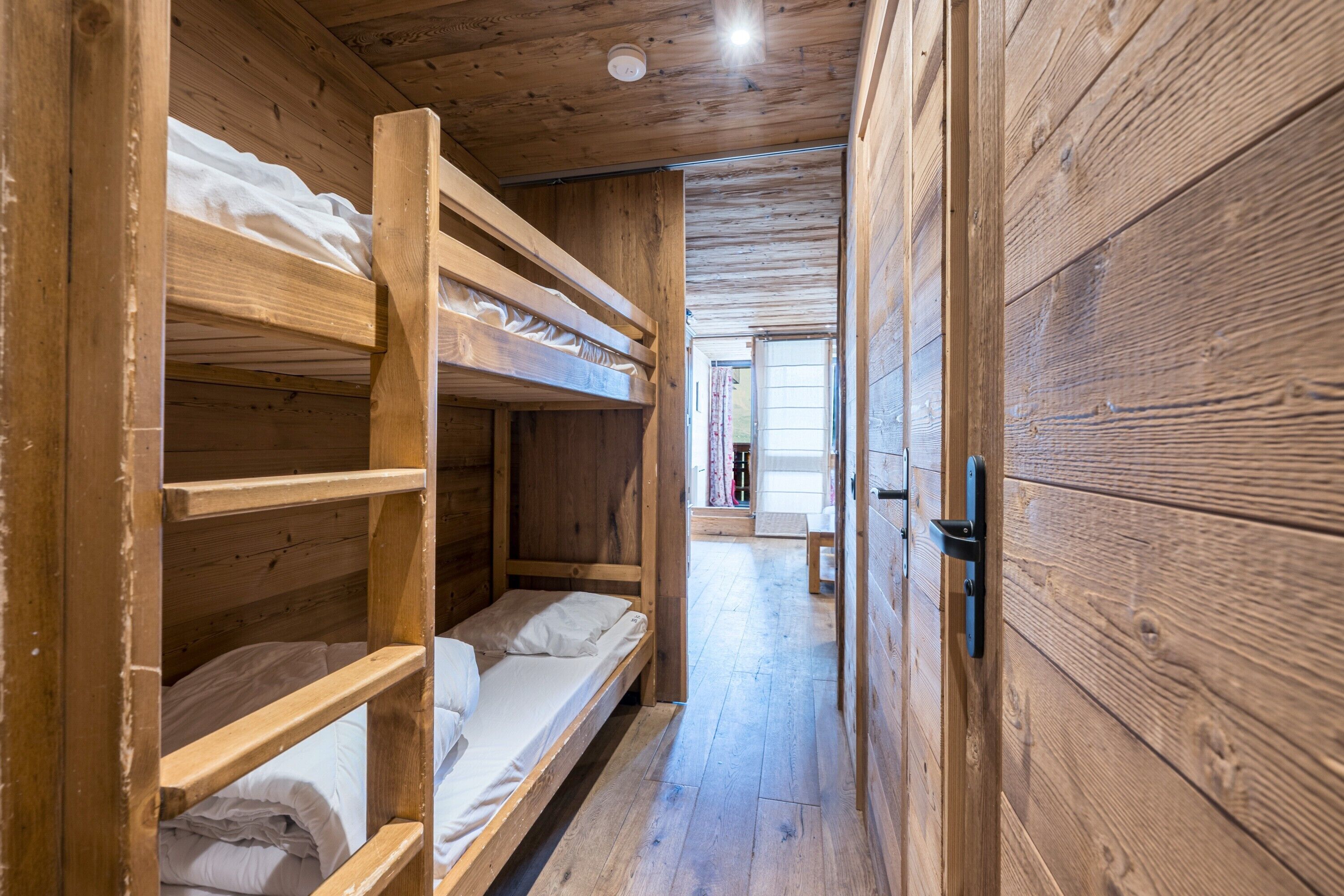 Een houten interieur met stapelbedden, een houten deur en een gang die leidt naar een kamer met een raam.