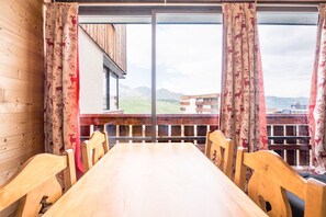 Un tavolo da pranzo in legno con sedie, vista sulle montagne attraverso ampie finestre e tende a motivi decorativi.