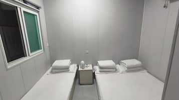 A small, single-room hotel room with two beds, a window, and a wall-mounted air conditioning unit.