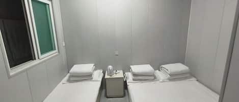 A small, single-room hotel room with two beds, a window, and a wall-mounted air conditioning unit.