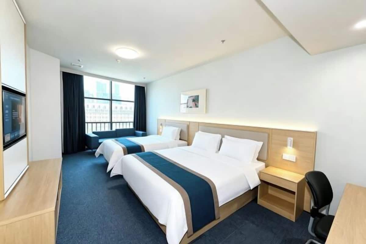 A hotel room with two beds, a desk, a chair, a TV, and a window with a view of buildings.