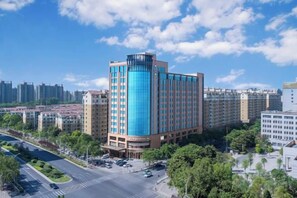 A multi-story hotel with a prominent blue glass facade, surrounded by other buildings and a road with vehicles. - Jinjiang Inn (Zhengzhou High-speed Railway East Station Zhengdong MixC Branch) (Zhengzhou)