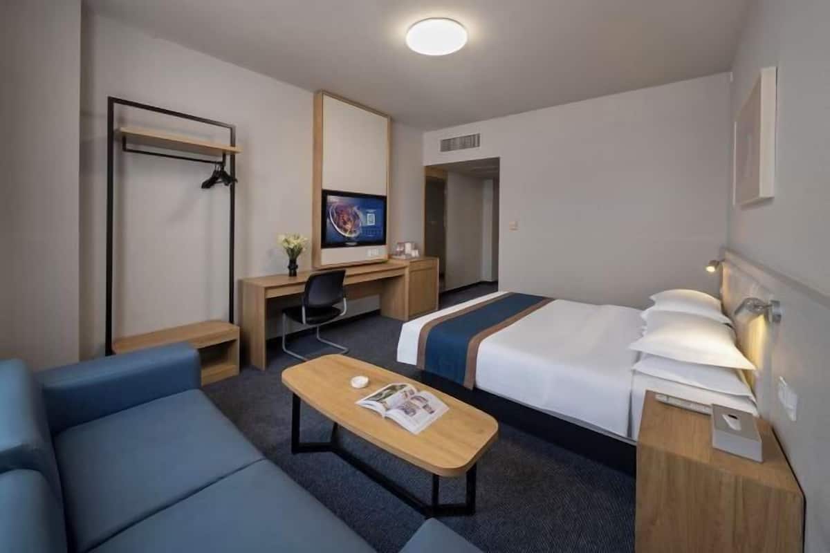 A hotel room with a bed, a desk, a chair, a small table with a magazine, a coat rack, and a mirror.
