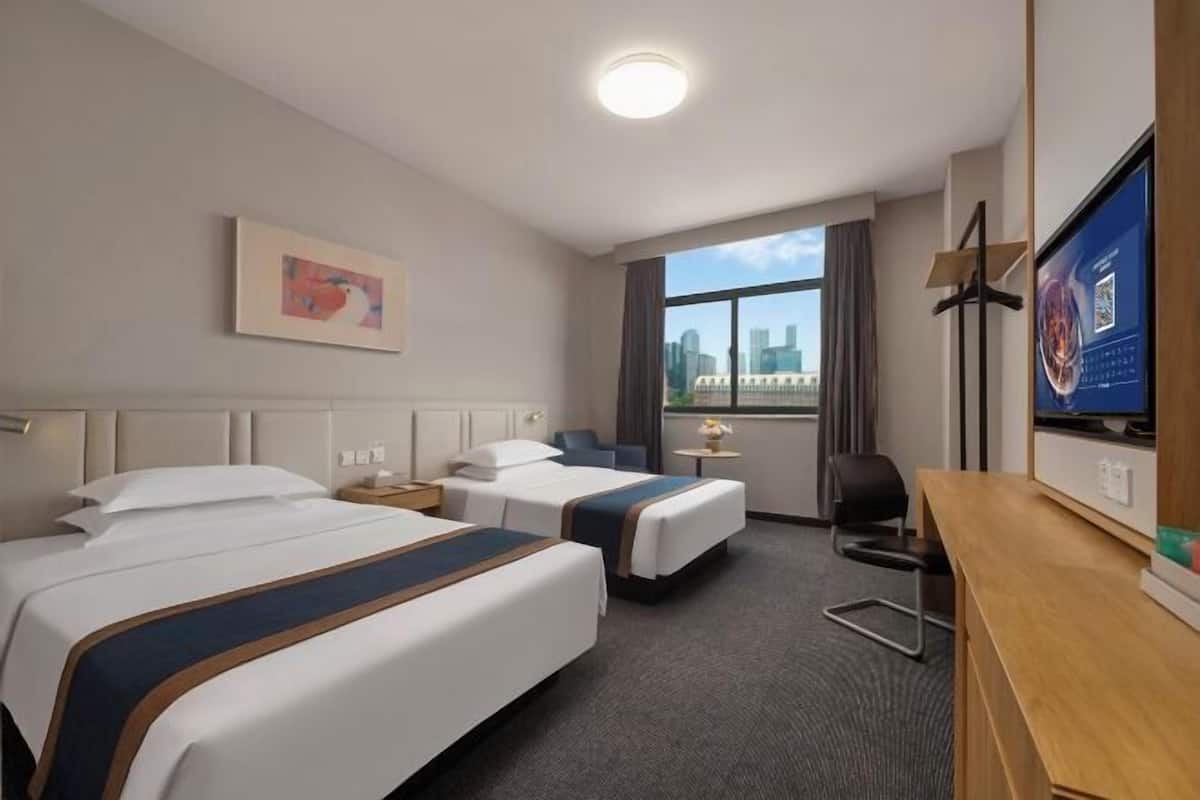 A hotel room with two beds, a desk, a TV, and a window with a city view.