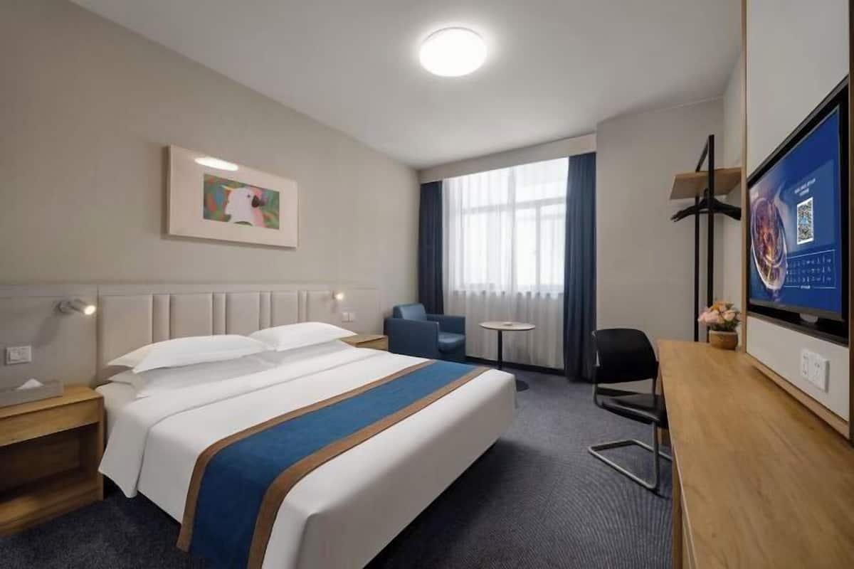 A hotel room with a bed, a desk, a chair, a small table, a TV, and a window with curtains.