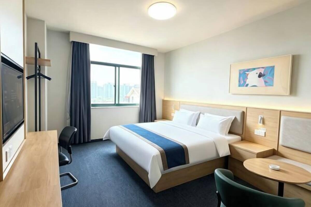 A hotel room with a bed, a desk, a chair, a small table, a TV, and a window with curtains.