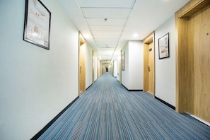 Un long couloir étroit, typique d’un hôtel, avec des portes en bois d’un côté et des tableaux encadrés de l’autre.