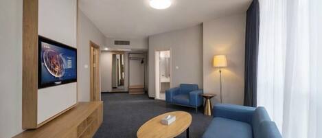 Habitación de hotel con un televisor de pantalla plana montado en una consola de madera, un sofá azul, una mesita redonda y una lámpara.