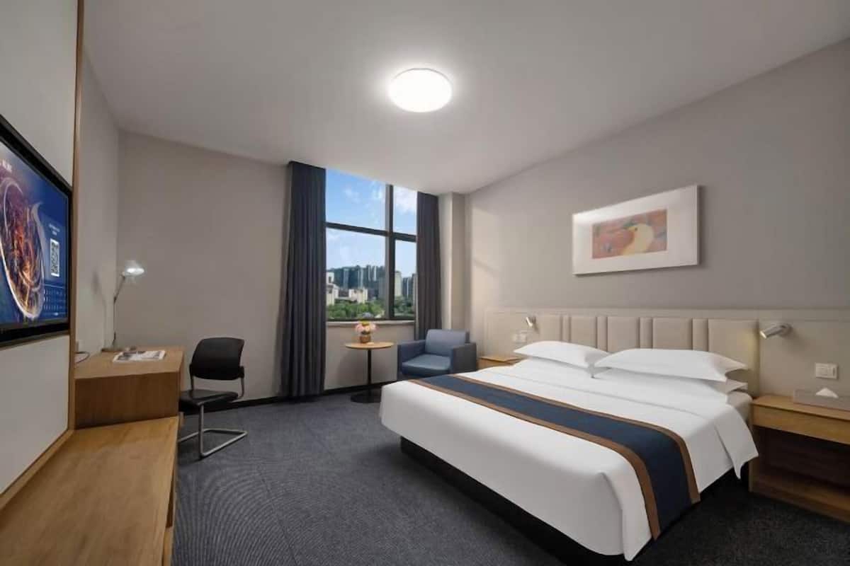 A hotel room with a large bed, a desk, a chair, a small table, and a window with a city view.
