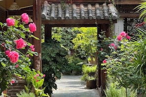 Un sendero en un jardín con una estructura de techo de tejas tradicionales, rodeado de exuberante follaje y plantas con flores.