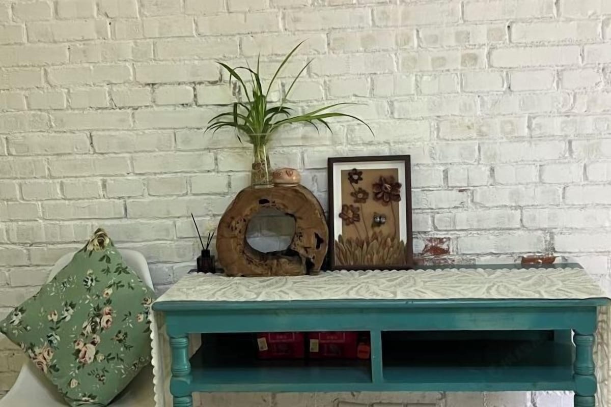 A teal-colored wooden console table with a decorative top, a potted plant, and a framed picture against a white brick wall.
