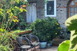 An outdoor seating area with wicker chairs, a potted plant with yellow flowers, and a brick wall with a window.