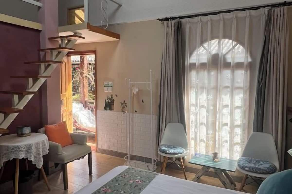Une pièce comprenant un lit, deux chaises, une petite table, un escalier et une fenêtre avec des rideaux.