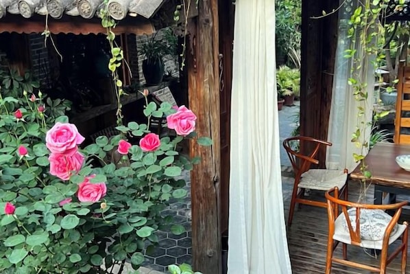 A wooden table and chairs outside a building with a tiled roof, surrounded by pink roses.