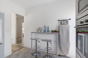 Una cocina moderna con horno de acero inoxidable, una mesita con dos taburetes y una tabla de planchar colgante.