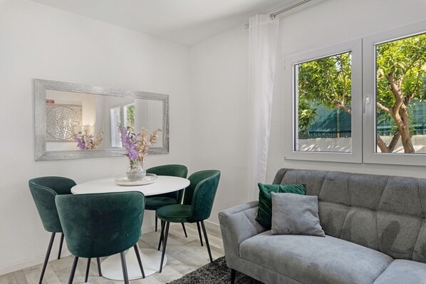 Una sala de estar moderna con un sofá gris, sillas verdes, una mesa blanca y un amplio ventanal con vista a un árbol.