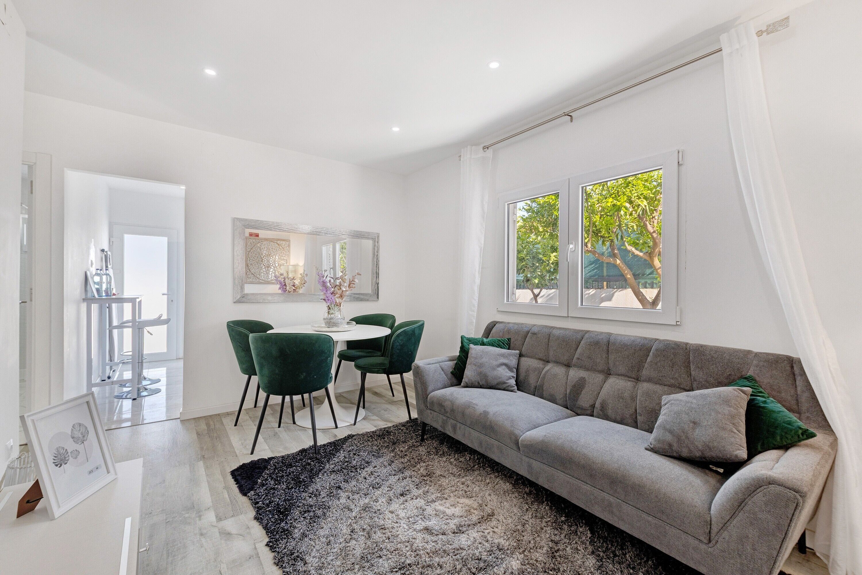 Uma sala de estar moderna com um sofá cinzento, poltronas verdes, uma mesa redonda e uma grande janela com vista para árvores.