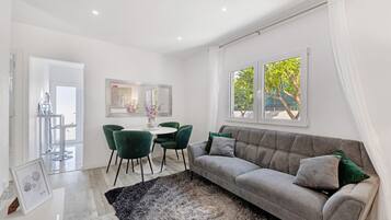 Uma sala de estar moderna com um sofá cinzento, poltronas verdes, uma mesa redonda e uma grande janela com vista para árvores.