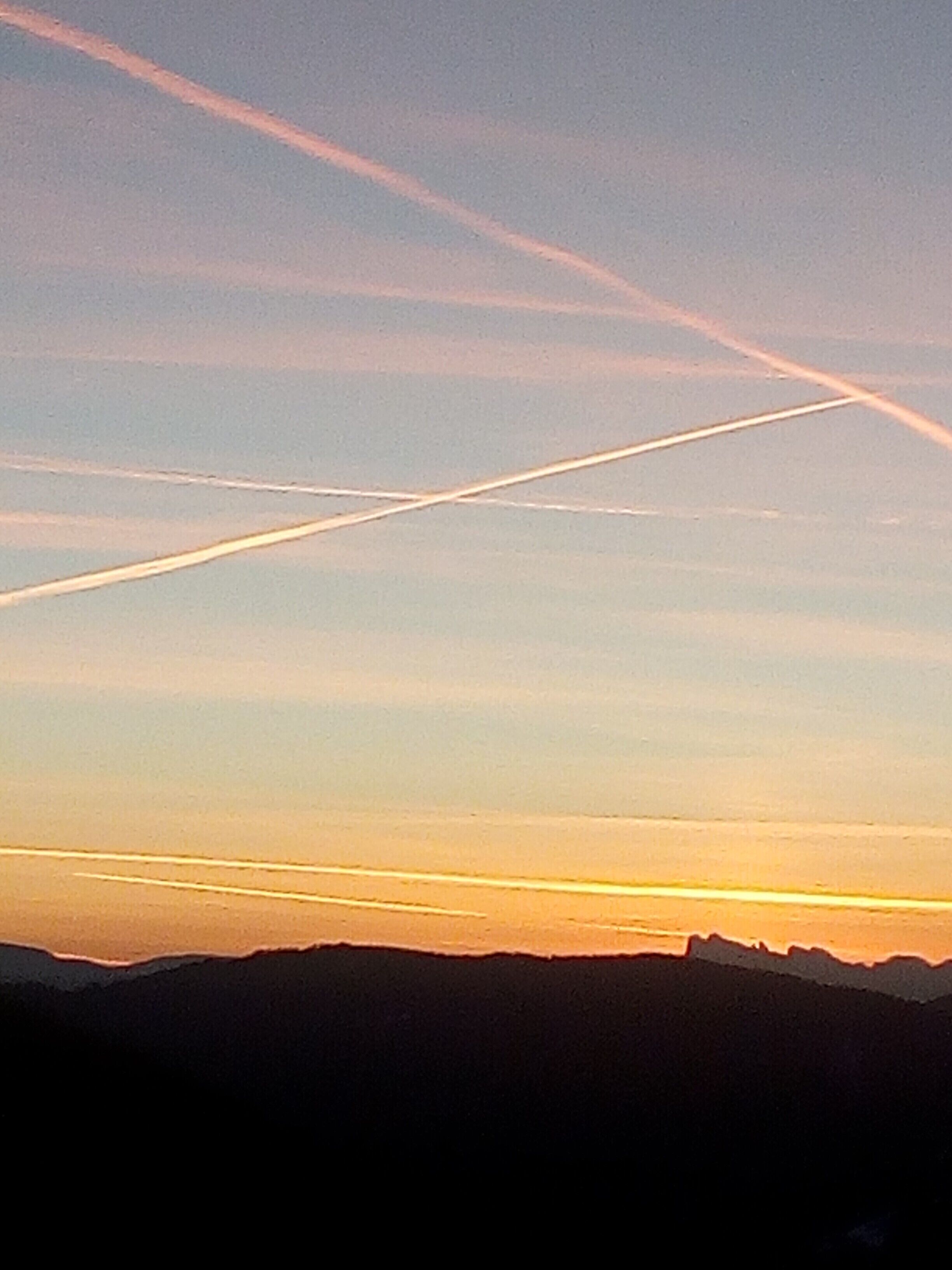 Een zonsondergang boven een bergachtig landschap, met zichtbare condensstrepen aan de lucht.