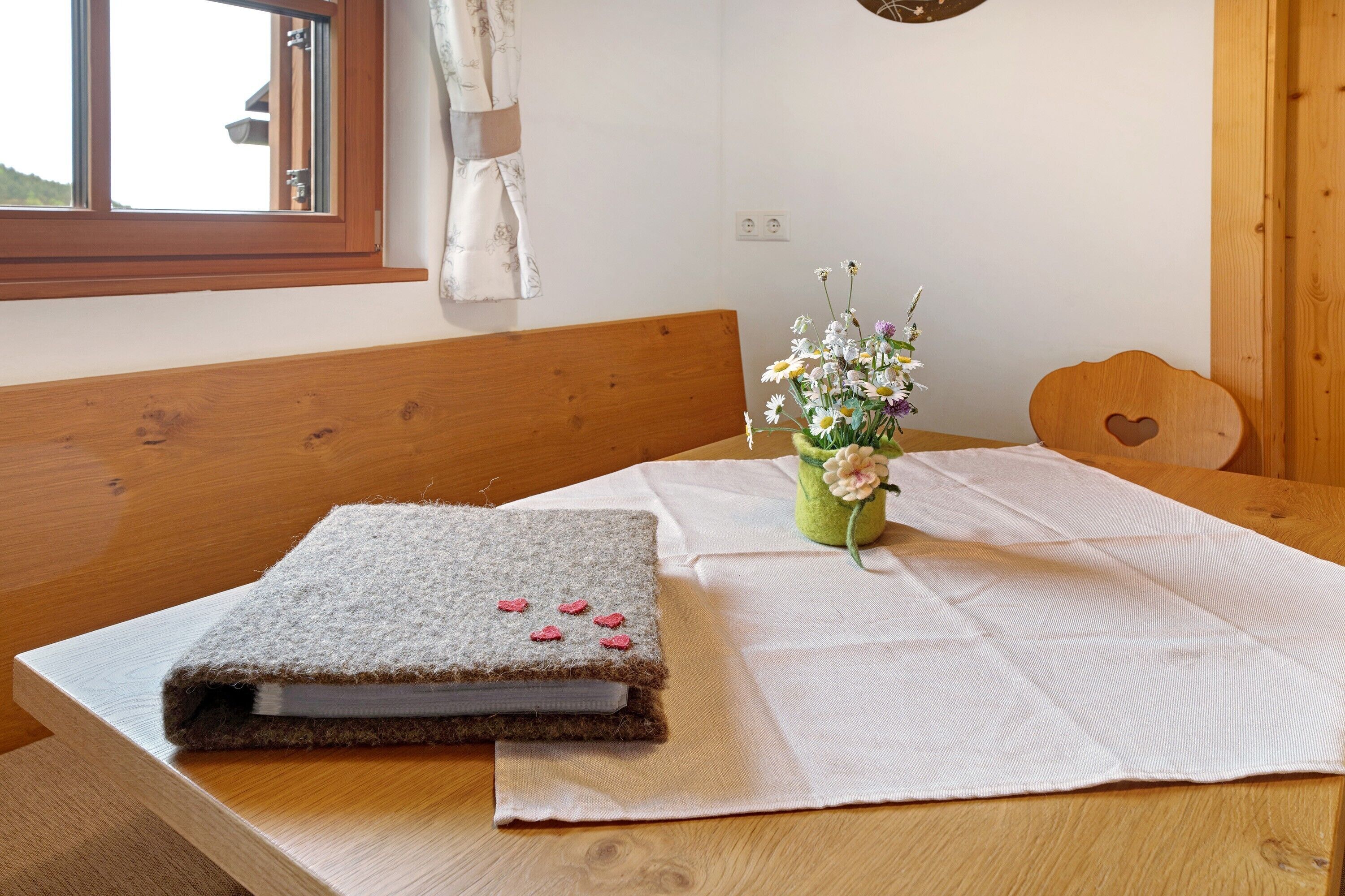 Une table en bois recouverte d’une nappe blanche, un banc, un petit vase avec des fleurs et un livre orné de fleurs roses.