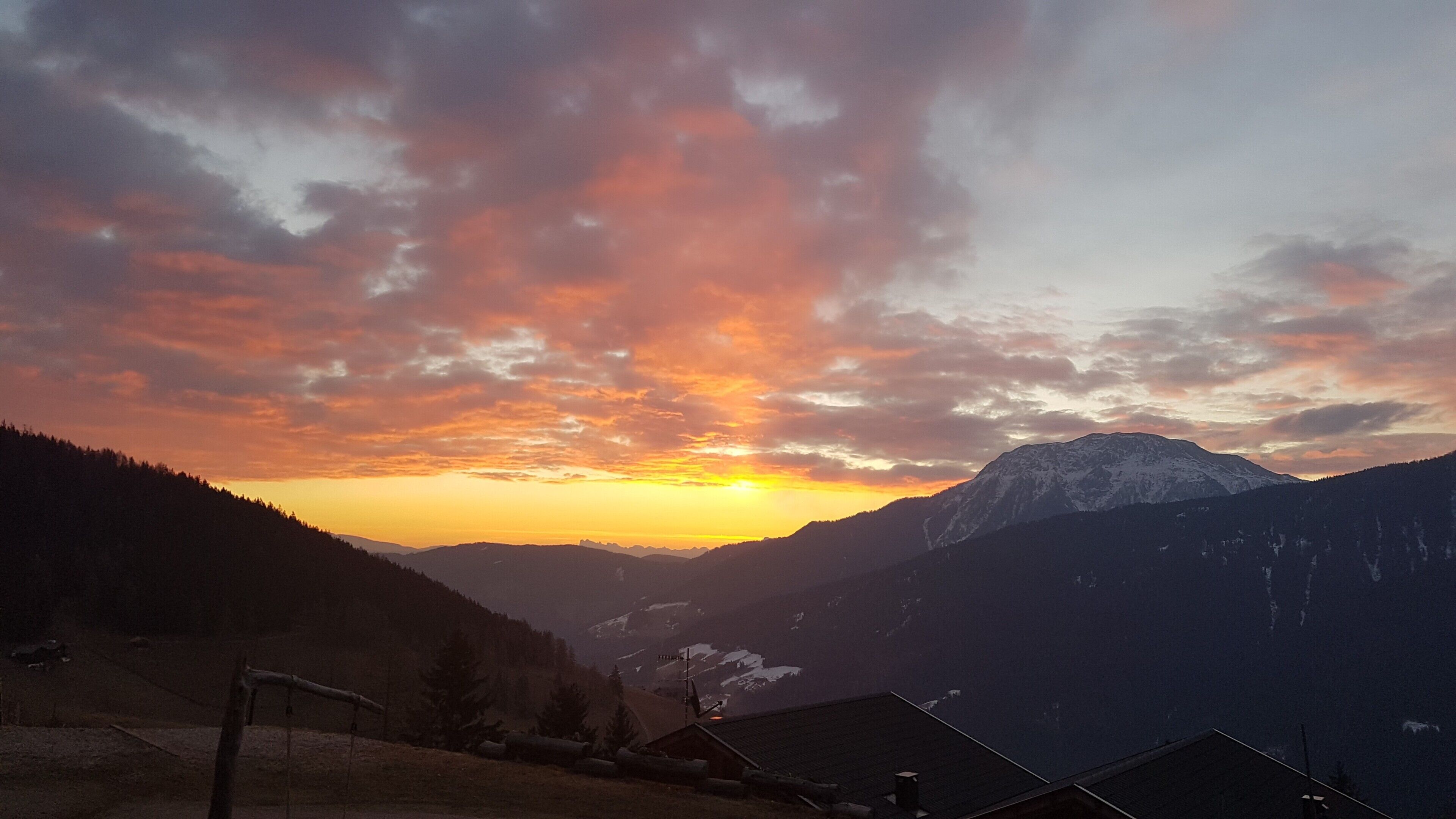 A mountain landscape with a sunset, snow-capped peaks, and a few buildings in the foreground.