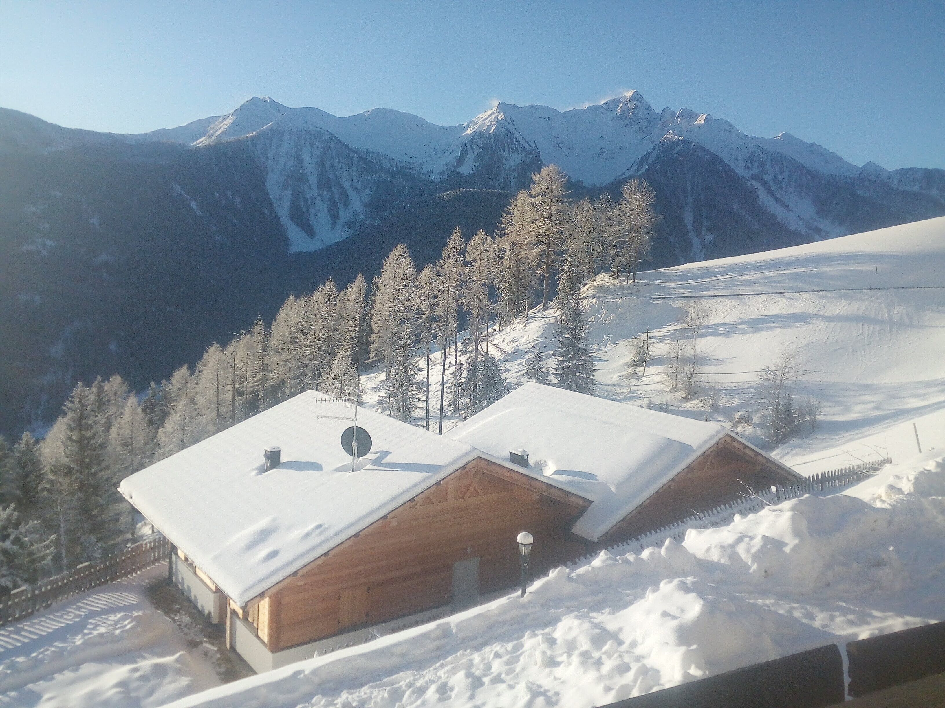 Une cabane en bois, dont le toit est recouvert de neige, entourée d’un paysage enneigé et avec des montagnes en arrière-plan.