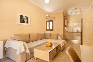 Un salon avec un canapé, une petite table sur laquelle se trouve un panier de fruits, un tableau encadré au mur, et une cuisine en arrière-plan.