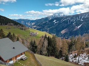 Un chalet de montagne avec une façade en bois, une terrasse couverte et une vue sur une vallée et des montagnes enneigées.
