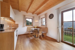A kitchen with wooden cabinets, a dining table with chairs, a window with curtains, and a view of a wooden fence and greenery outside.