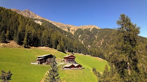 Un albergue de montaña con dos edificios principales, rodeado por un valle verde exuberante y un bosque denso.