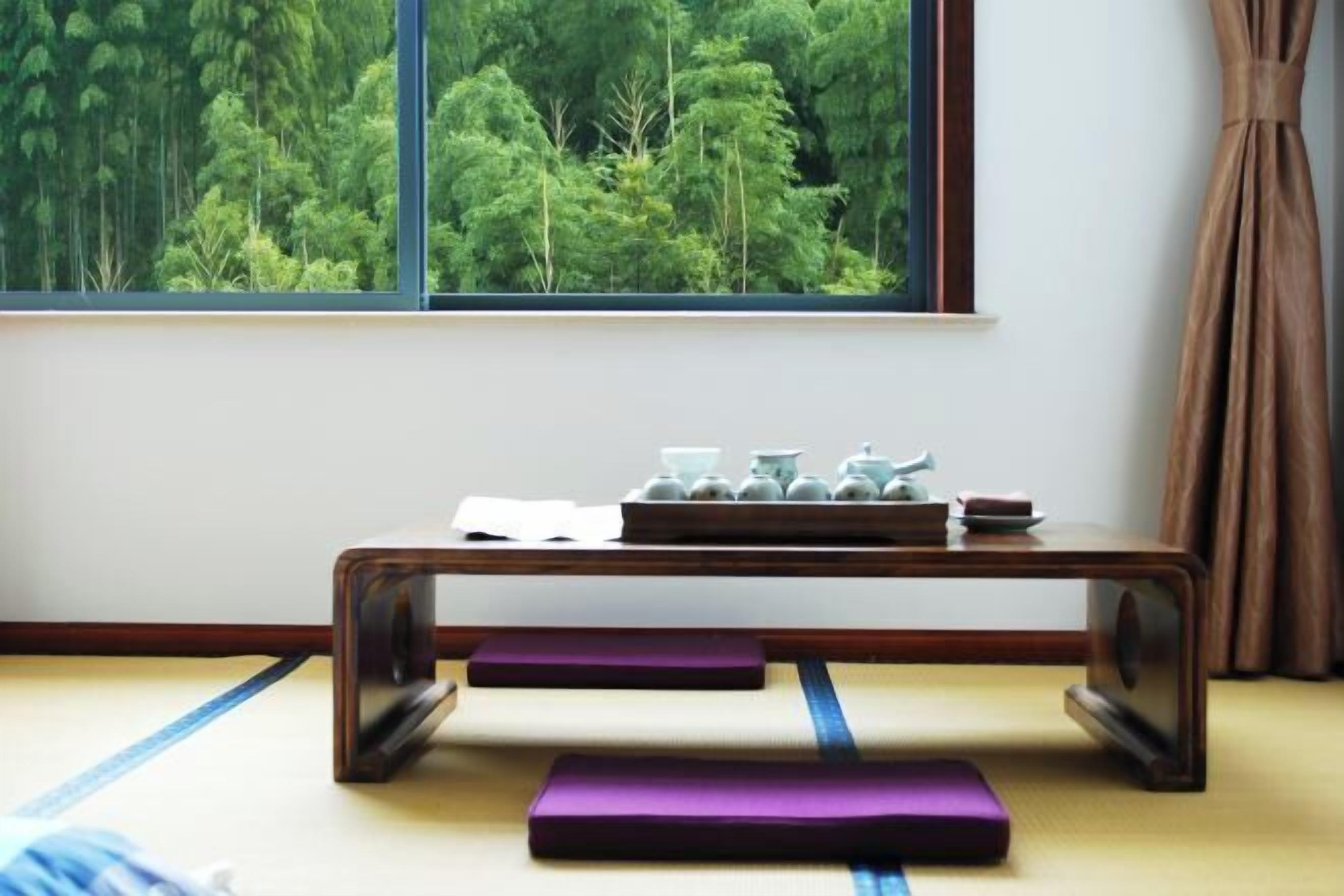 Ein Raum mit Tatami-Matten, einem niedrigen Holztisch, einem Fenster mit Blick auf einen Wald und einem Vorhang.