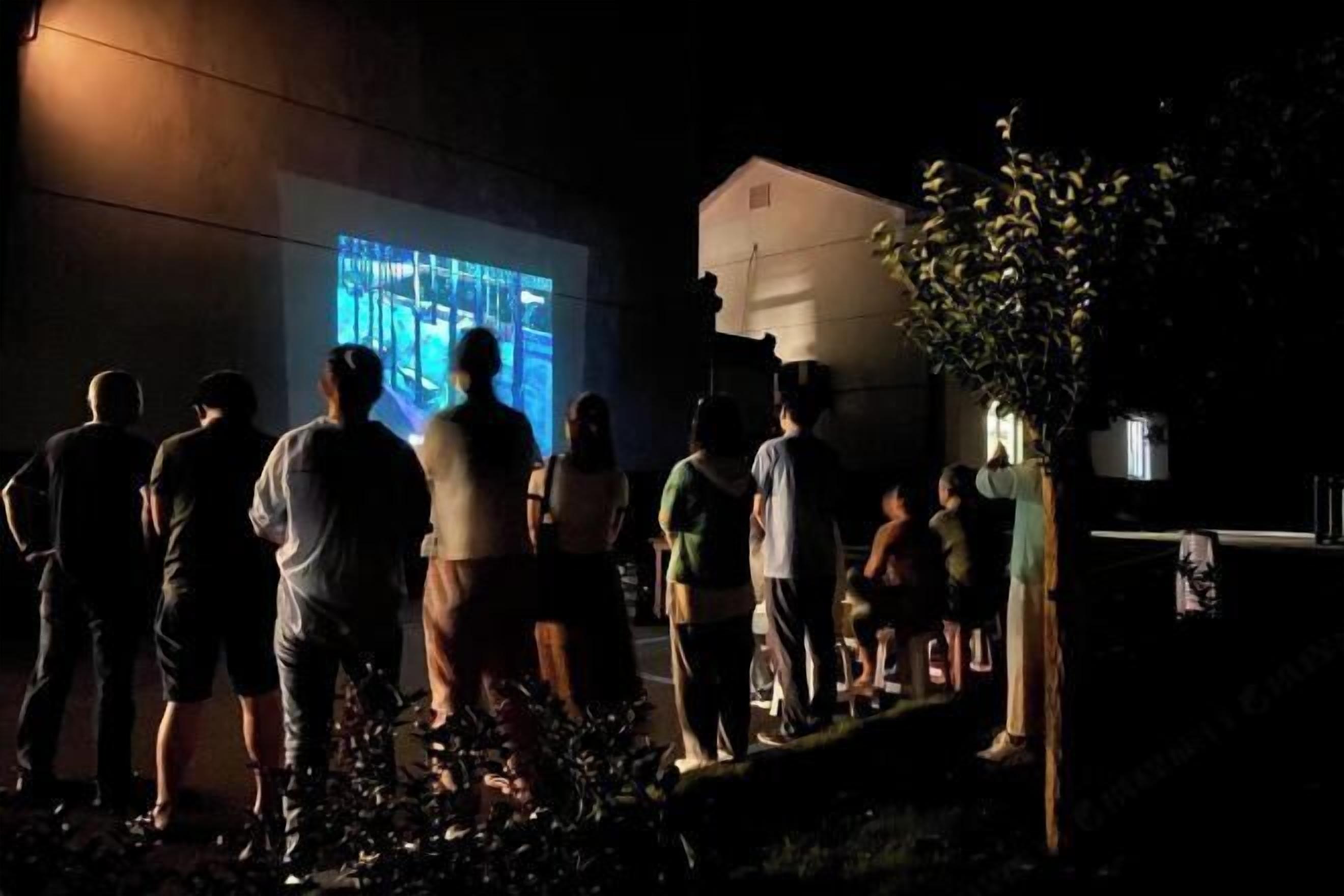 A group of people gathered outdoors at night, watching a large screen displaying an image.