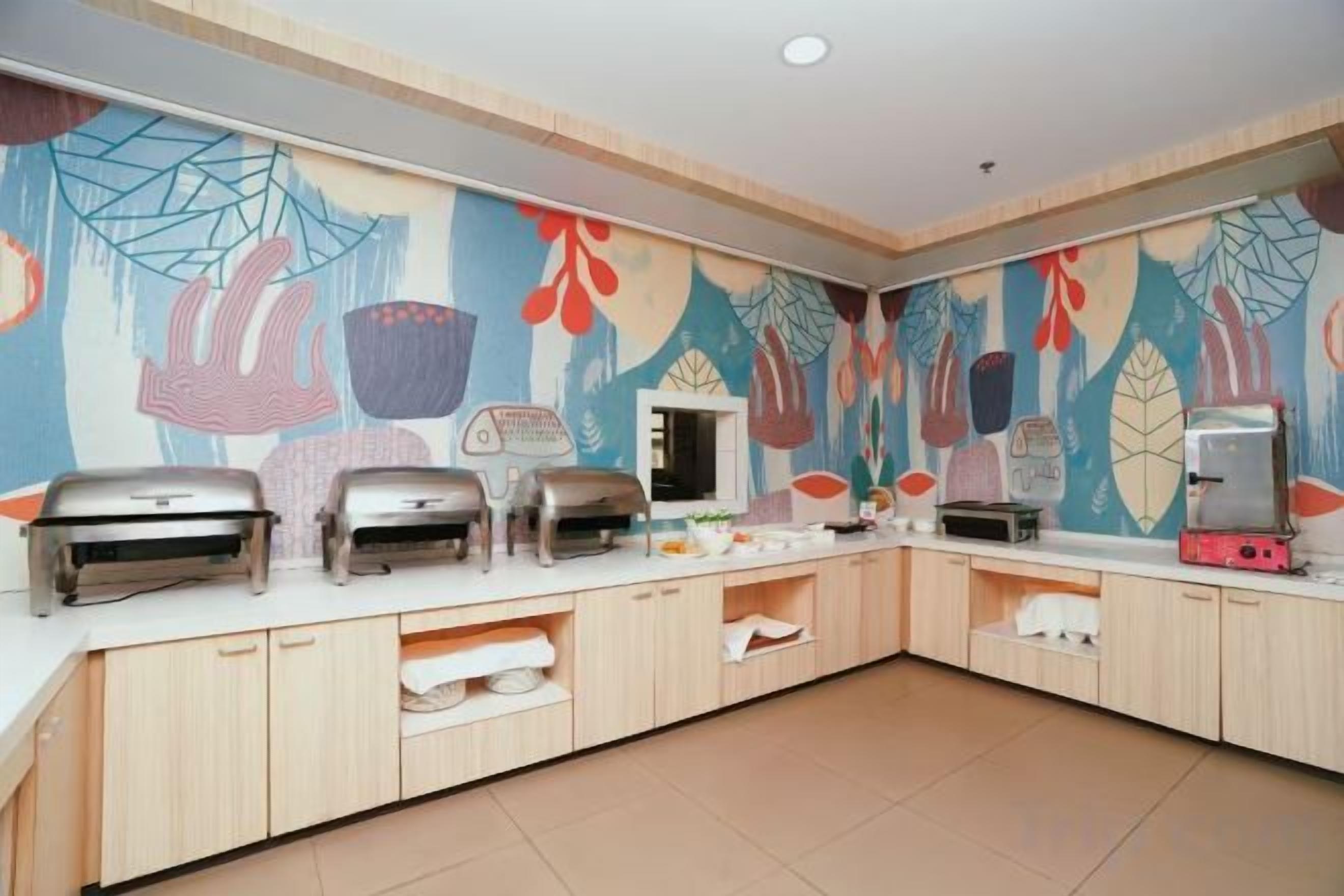 A buffet setup with multiple grills and a refrigerator, a tiled floor, and a wall with a colorful mural.