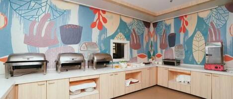 Disposición de un bufé con varias parrillas y un refrigerador, piso de baldosas y una pared con un mural colorido.