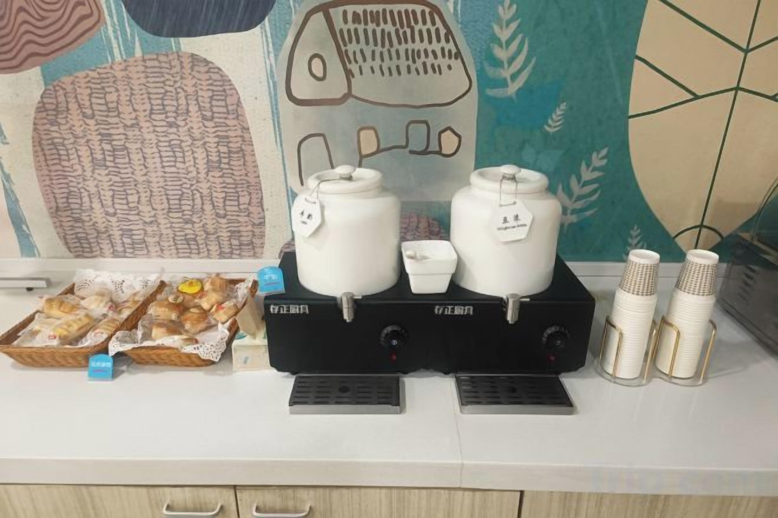 Uma bancada de cozinha com duas cafeteiras, uma cesta de doces e uma pilha de copos descartáveis.
