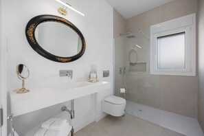 A modern bathroom with a large oval mirror, a white sink, a toilet, and a glass-enclosed shower.