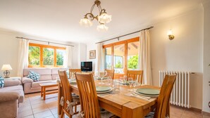 A dining area with a wooden table set for six, surrounded by chairs, a sofa, and a television in the background.