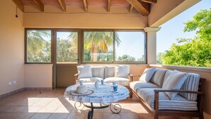A sunlit patio with a round table, striped sofa, and a chair, featuring large windows and a view of greenery.