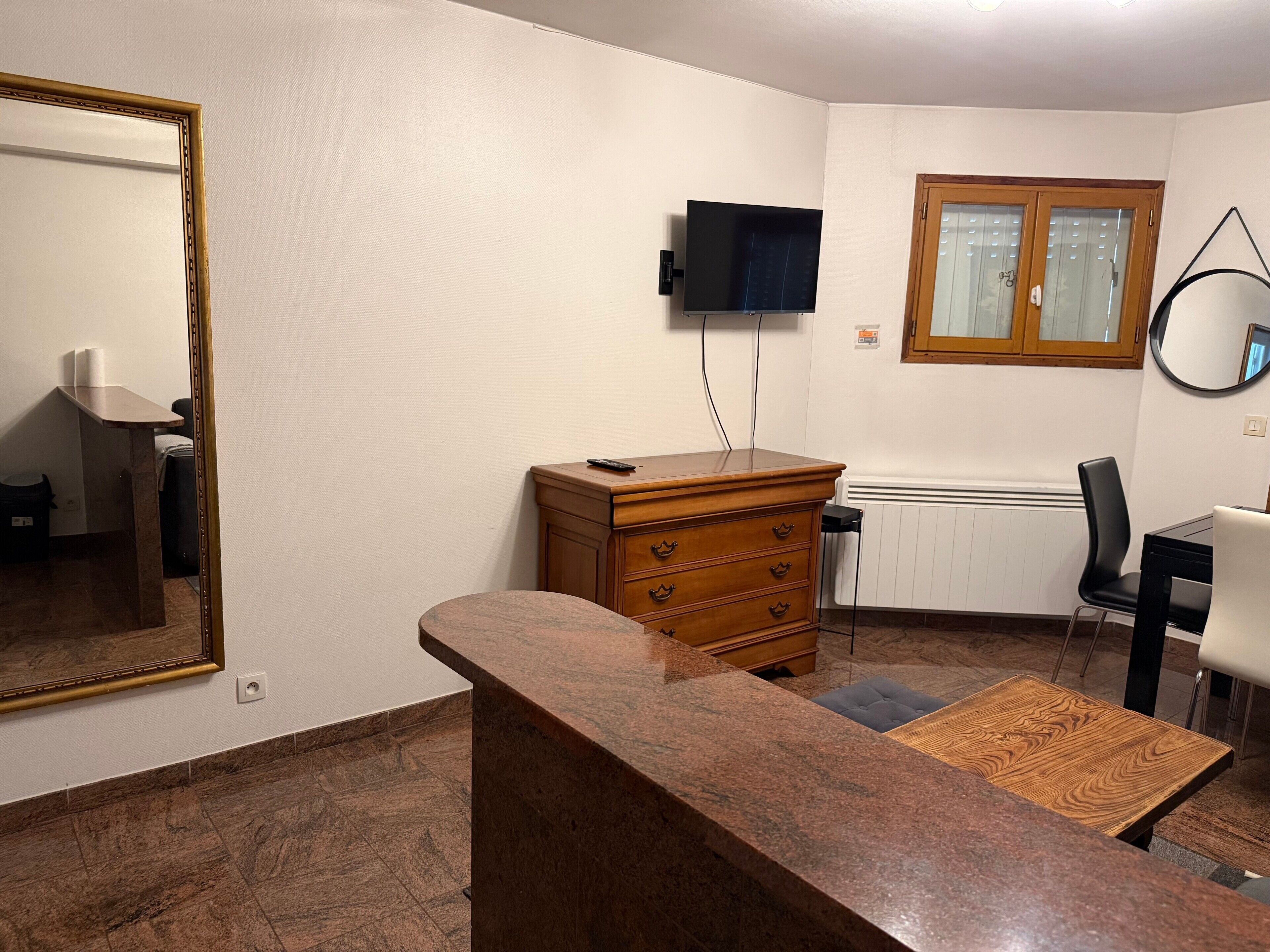 A room with a wooden dresser, a television mounted on the wall, a mirror, and a window with blinds.