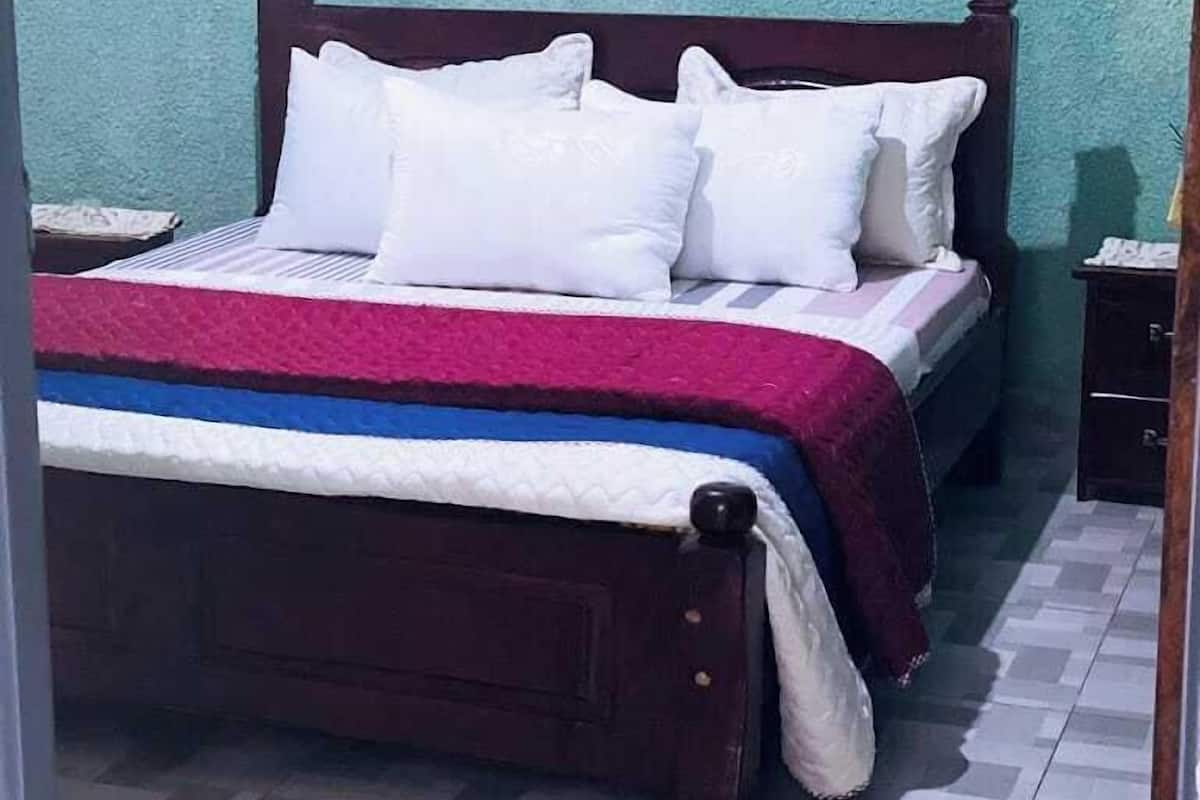 Una cama con almohadas blancas y una colcha a rayas rojas y blancas, un cabecero de madera y un piso de baldosas.