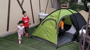 Una tenda verde montata su erba sintetica, con un lavandino e una sedia nelle vicinanze.