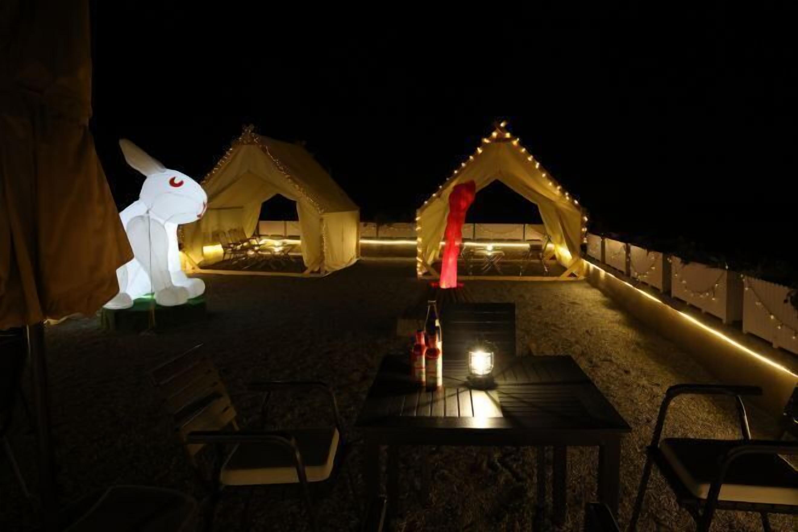 Un espace extérieur aménagé pour les soirées, avec des tables et des chaises, illuminé par des guirlandes lumineuses et comprenant deux tentes avec une lumière rouge au centre.