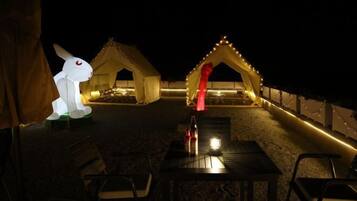 Un espace extérieur aménagé pour les soirées, avec des tables et des chaises, illuminé par des guirlandes lumineuses et comprenant deux tentes avec une lumière rouge au centre.