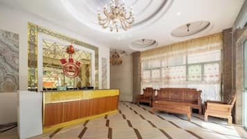 A hotel lobby with a reception desk, a chandelier, a large window with curtains, and a seating area with wooden furniture.