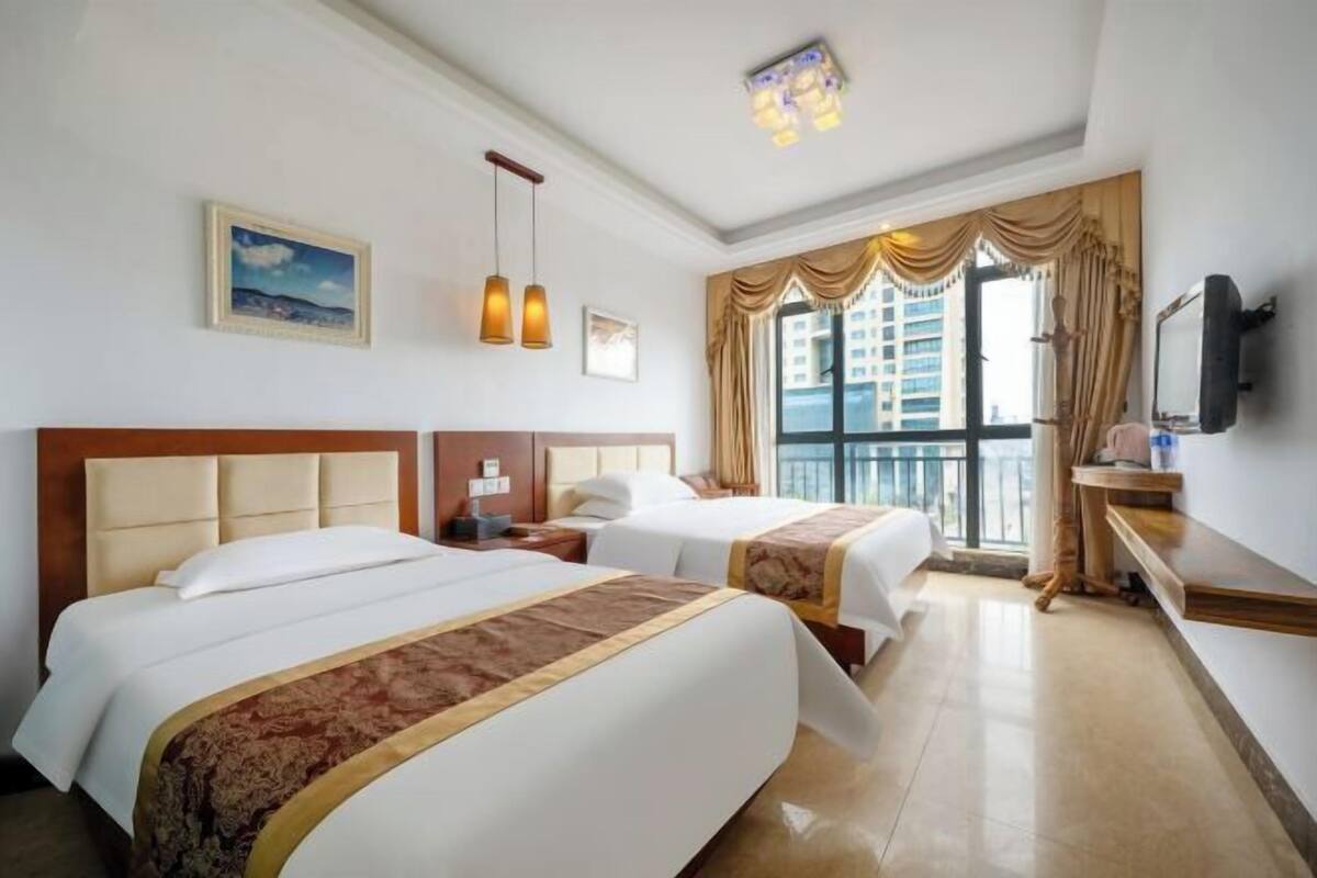 Een hotelkamer met twee bedden, een groot raam met gordijnen, een flatscreen televisie en een plafondlamp.